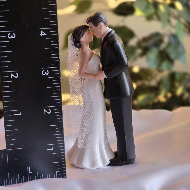 May include: A wedding cake topper featuring a bride and groom figurine. The bride wears a white dress and veil, while the groom is in a black tuxedo. The figures are approximately 3 inches tall.