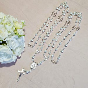 May include: Two pearl rosaries with silver-tone crucifixes and oval pendants featuring religious imagery. The rosaries are draped on a light pink surface, with a bouquet of white flowers in the background.
