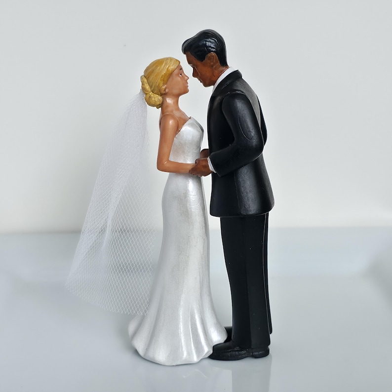 May include: A wedding cake topper featuring a bride and groom figurine. The bride wears a white strapless gown and a veil. The groom is in a black tuxedo. The figurines are facing each other, holding hands, and are close together.
