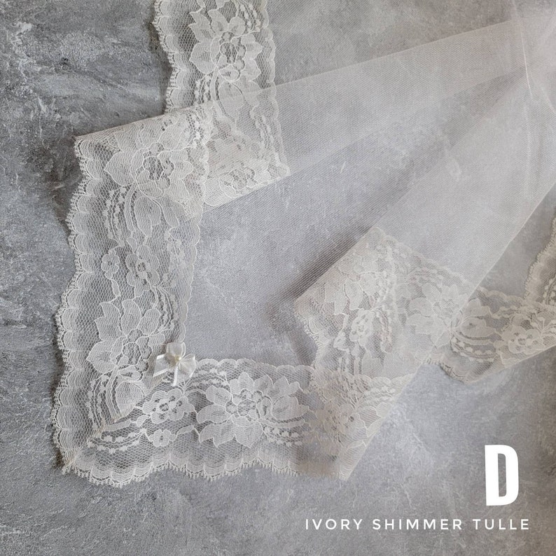 Puede incluir: Una tela de tul color marfil brillante con un borde de encaje floral ancho e intrincado en los extremos. Un peque&ntilde;o y delicado lazo de cinta color marfil est&aacute; sujeto al borde de encaje. El texto D IVORY SHIMMER TULLE aparece en la esquina inferior derecha.