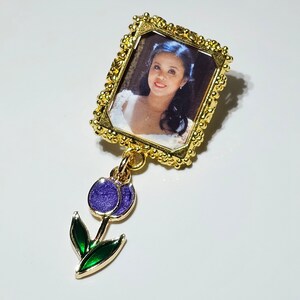 May include: A gold-toned picture frame with a photo of a woman with long brown hair. The frame is attached to a purple and green enamel flower charm.