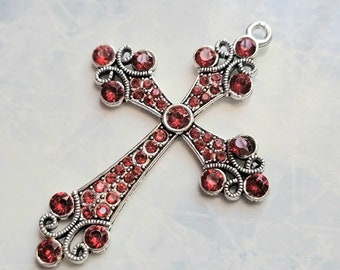 Op de afbeelding: Een zilveren kruis hanger met een gedetailleerd ontwerp en rode strass steentjes.