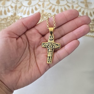Puede incluir: Un collar con colgante de cruz de color dorado. La cruz presenta un relieve detallado de una figura religiosa y está suspendida de una delicada cadena. El colgante mide aproximadamente 5 cm de largo.