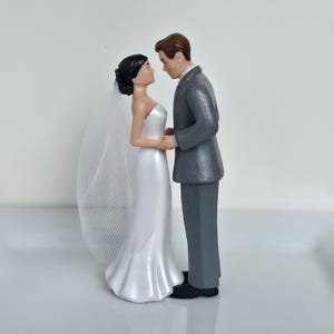 May include: A wedding cake topper featuring a bride and groom figurine. The bride wears a white, strapless gown and veil, while the groom is in a gray suit. They are holding hands and looking at each other.
