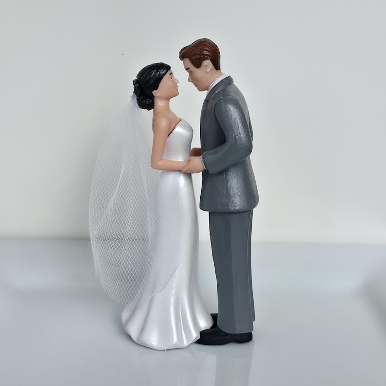 May include: A wedding cake topper featuring a bride and groom figurine. The bride wears a white, strapless gown and veil, while the groom is in a gray suit. They are holding hands and looking at each other.
