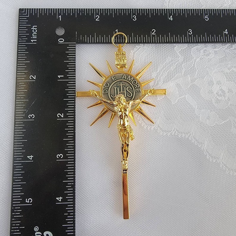 May include: A gold-colored crucifix with a sunburst design behind the figure of Jesus. The cross is approximately 4 inches long. The center medallion reads "DIO E AMORE" and "IHS".