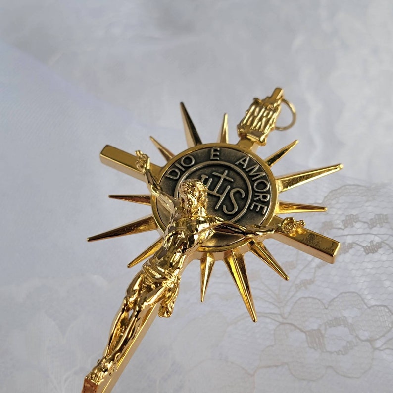 May include: A gold-colored crucifix with a detailed depiction of Jesus Christ. The cross features a sunburst design behind the figure, with the inscription "DIO E AMORE" and the letters "IHS". The crucifix is likely a religious item.