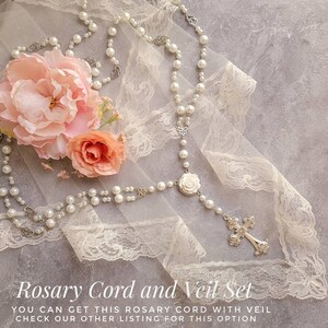 May include: A white pearl rosary cord with a silver cross and a white rose charm, displayed on a white lace veil with pink flowers.
