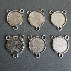 May include: Six silver-toned metal round pendant trays with three loops on each. The trays have a smooth, slightly textured surface. One tray has a decorative swirl pattern.
