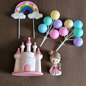 May include: A set of cake toppers featuring a pink castle, a rainbow with clouds, a princess doll, and a cluster of pastel-colored balloons.