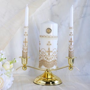 Puede incluir: Un juego de dos velas de unidad de boda blancas con bordados dorados y un soporte de metal dorado. La vela más grande tiene un pequeño adorno dorado en la parte delantera.