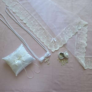 May include: A collection of white religious items, including a satin ring pillow with lace and a bow, two necklaces with cross pendants, a lace-trimmed veil, and a small heart-shaped box with coins. The items are arranged on a pink surface.
