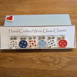 May include: A set of six circular wine glass charms in a white box labeled Hand Crafted Wine Glass Charms. Each charm features a button-like design with various colored polka dots in red, blue, and light blue, attached to silver-toned metal rings.