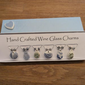 May include: A set of six hand crafted wine glass charms in a white box labeled Hand Crafted Wine Glass Charms. Each charm features a unique ceramic pendant in blue, white, and green tones with silver metal rings and small decorative beads.