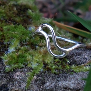 Pode incluir: Um anel prateado com um design abstrato e fluido. O anel está colocado sobre um fundo de musgo verde e uma superfície de pedra, com fios de erva em primeiro plano. O design do anel apresenta uma forma contínua e em laço.