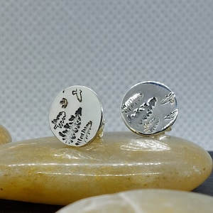 May include: A pair of silver earrings with a fern design. The earrings are round and have a textured surface.