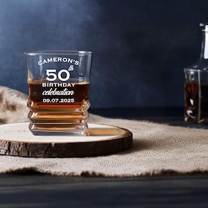 May include: A whiskey glass with the text "Cameron's 50th Birthday Celebration 09.07.2025" is filled with amber liquid, sitting on a wooden coaster. A decanter is in the background. The glass has a unique, ridged design.