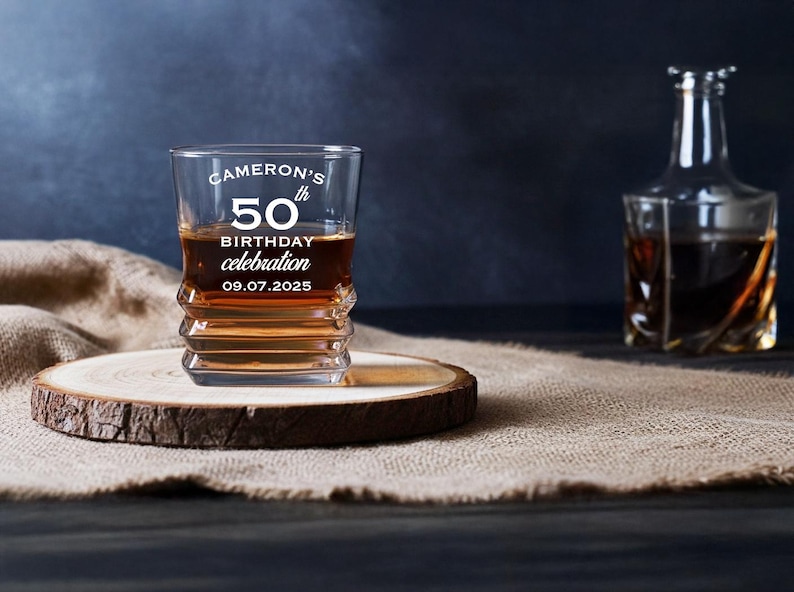 May include: A whiskey glass with the text "Cameron's 50th Birthday Celebration 09.07.2025" is filled with amber liquid, sitting on a wooden coaster. A decanter is in the background. The glass has a unique, ridged design.