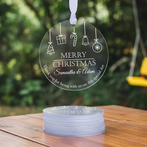 May include: A circular clear acrylic ornament hanging by a white ribbon, featuring white line art of a Christmas tree, gift, candy cane, bell, and bauble. It reads MERRY CHRISTMAS, Samantha & Adam, and Thank you for being with us on this special day.