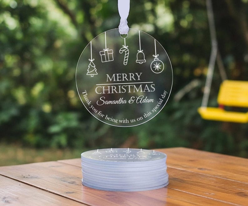 May include: A circular clear acrylic ornament hanging by a white ribbon, featuring white line art of a Christmas tree, gift, candy cane, bell, and bauble. It reads MERRY CHRISTMAS, Samantha & Adam, and Thank you for being with us on this special day.