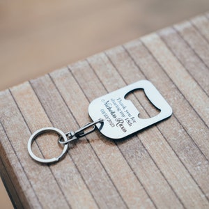 A single personalized stainless steel bottle opener keychain lying on a wooden surface, highlighting the engraved text and the metal’s reflective finish.