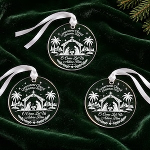 May include: Three circular clear acrylic ornaments featuring a white etched nativity scene with palm trees and a star. Each ornament displays the text Cornerstone Church Christmas 2026 and O Come Let Us Adore Him, finished with a white satin ribbon loop.