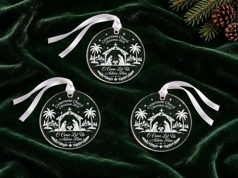 May include: Three circular clear acrylic ornaments featuring a white etched nativity scene with palm trees and a star. Each ornament displays the text Cornerstone Church Christmas 2026 and O Come Let Us Adore Him, finished with a white satin ribbon loop.