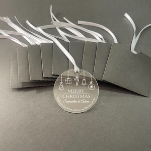 May include: A clear circular acrylic ornament featuring white etched Christmas icons, the text MERRY CHRISTMAS, Samantha & Adam, and Thank you for being with us on this special day. It includes a white ribbon and is paired with several dark gray tags.