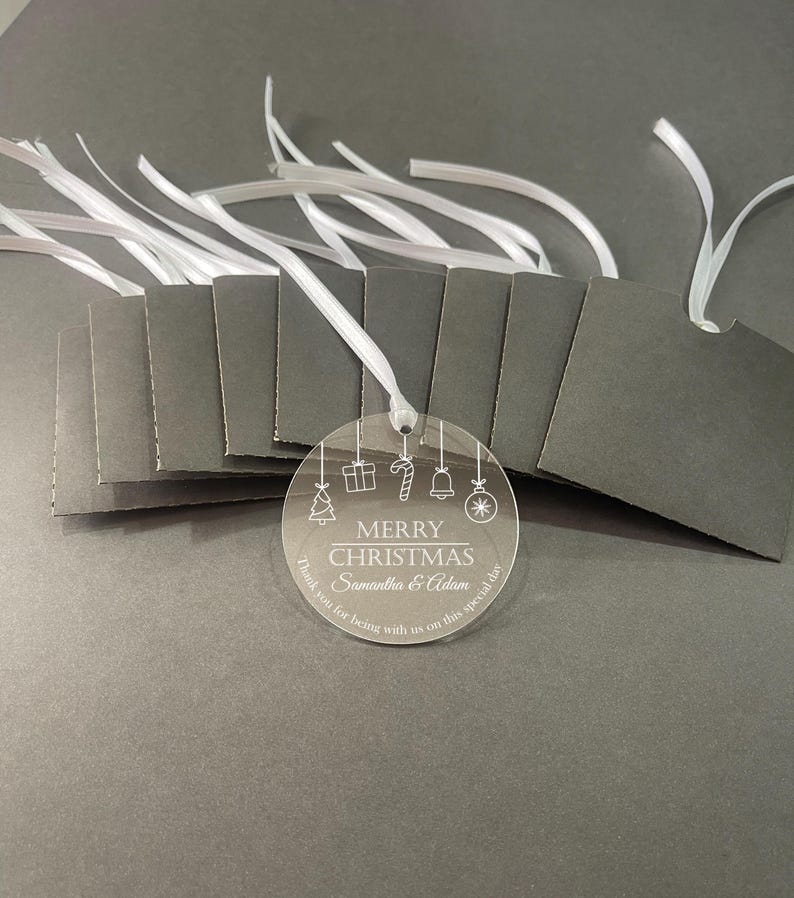 May include: A clear circular acrylic ornament featuring white etched Christmas icons, the text MERRY CHRISTMAS, Samantha & Adam, and Thank you for being with us on this special day. It includes a white ribbon and is paired with several dark gray tags.