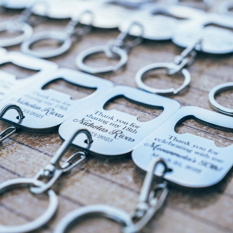 A close-up of multiple stainless steel bottle opener keychains arranged in rows, each engraved with personalized thank you messages. The polished metal finish shines under the light.