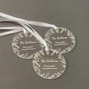 Three round acrylic ornaments with white ribbons are displayed on a dark background. Each ornament is engraved with “The BoHome” and a holiday message, surrounded by a wreath-like decorative border.
