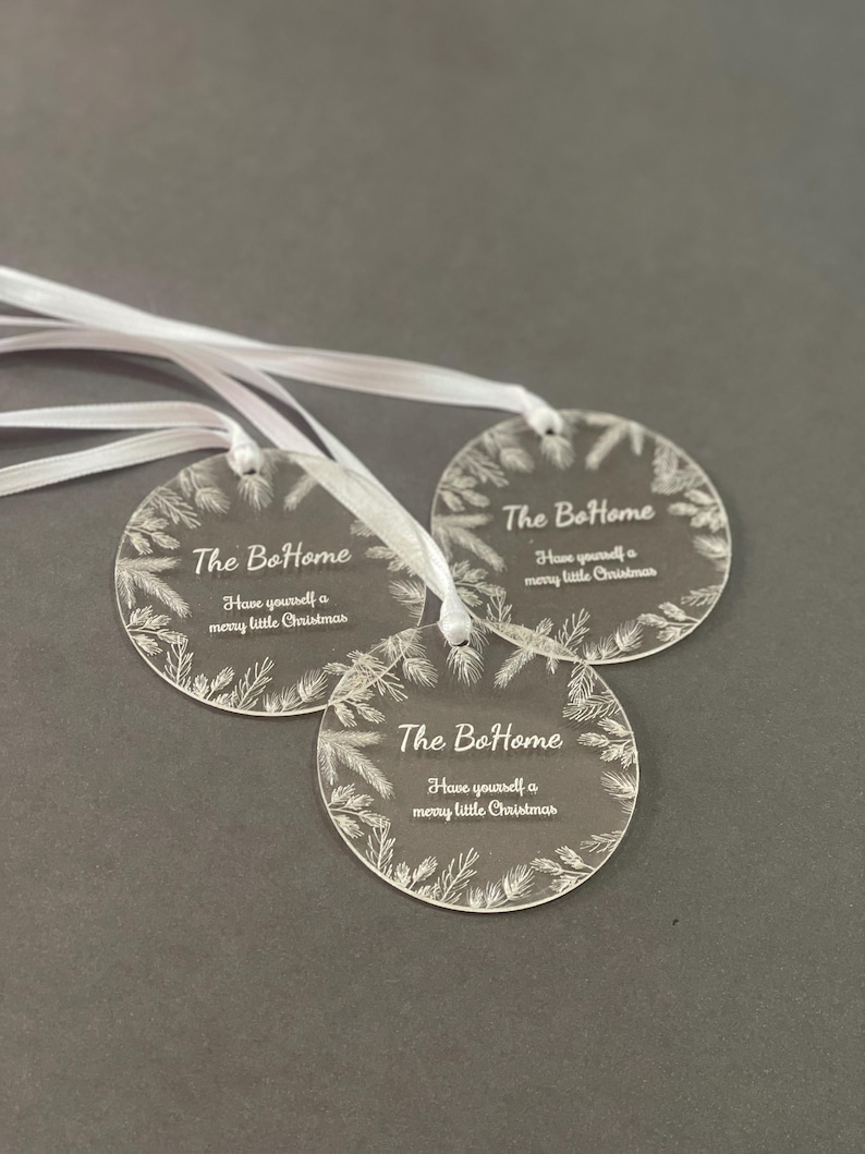 Three round acrylic ornaments with white ribbons are displayed on a dark background. Each ornament is engraved with “The BoHome” and a holiday message, surrounded by a wreath-like decorative border.