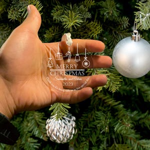 Hand holding a personalized acrylic Christmas ornament featuring a festive design, hanging from a tree with holiday decorations.