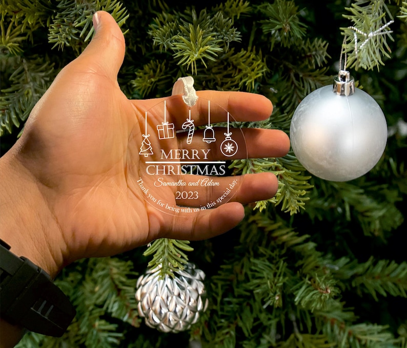 Hand holding a personalized acrylic Christmas ornament featuring a festive design, hanging from a tree with holiday decorations.