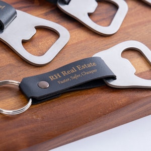Several bottle opener keychains with black leather wraps, each engraved with different personalized messages. The keychains are arranged on a wooden surface, showcasing the variety of engraving options.
