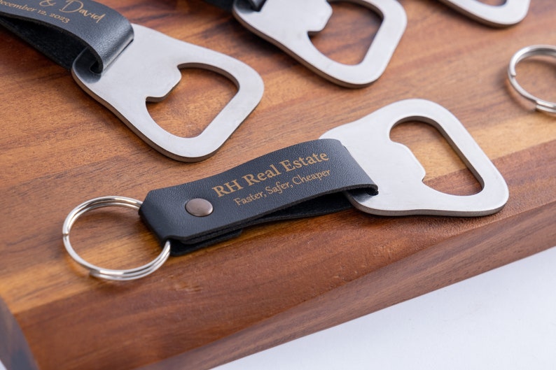 Several bottle opener keychains with black leather wraps, each engraved with different personalized messages. The keychains are arranged on a wooden surface, showcasing the variety of engraving options.