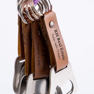 A bunch of leather-wrapped bottle opener keychains with brown leather wraps, hanging from a metal ring. The engraving on the leather is visible, showcasing the product’s portability.