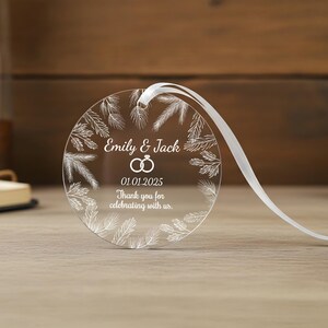 May include: A circular clear acrylic ornament featuring a white printed wreath of pine needles. The center displays the text Emily & Jack, a graphic of two interlocking rings, the date 01.01.2025, and the phrase Thank you for celebrating with us.