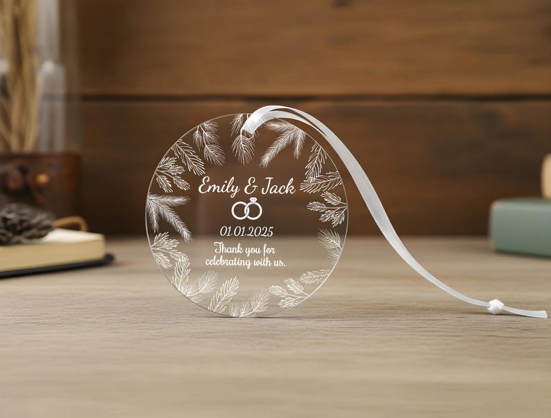 May include: A circular clear acrylic ornament featuring a white printed wreath of pine needles. The center displays the text Emily & Jack, a graphic of two interlocking rings, the date 01.01.2025, and the phrase Thank you for celebrating with us.