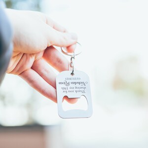 A hand holding a personalized stainless steel bottle opener keychain against a soft outdoor background. The engraving reads “Thank you for celebrating with me,” personalized with a name and date.