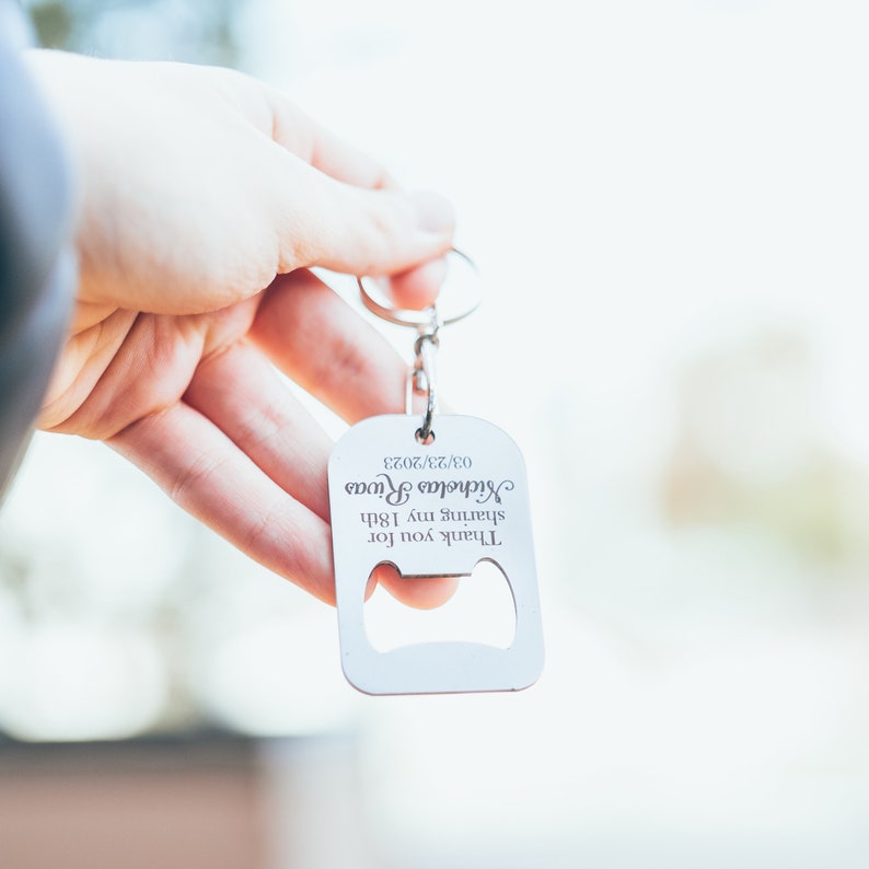 A hand holding a personalized stainless steel bottle opener keychain against a soft outdoor background. The engraving reads “Thank you for celebrating with me,” personalized with a name and date.