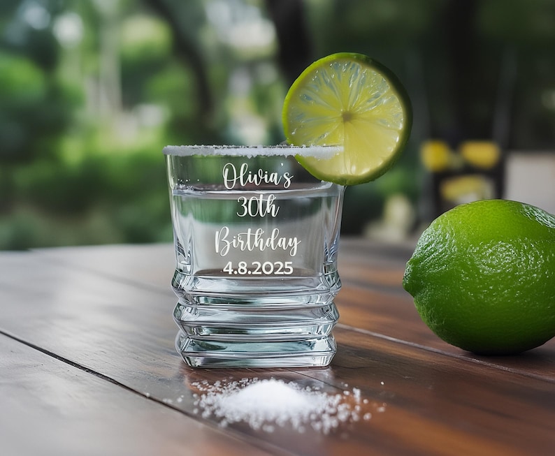 May include: A clear shot glass with a salt-rimmed edge, filled with liquid and a lime wedge. The glass is engraved with "Olivia's 30th Birthday 4.8.2025". A whole lime and a pile of salt are on the wooden surface.