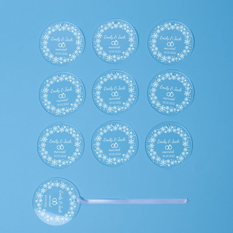 Ten round acrylic ornaments with a consistent design are laid out on a blue background. Each ornament is engraved with the names “Emily & Wade” and a holiday message, with one ornament featuring a ribbon attached for hanging.