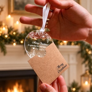 May include: A circular clear acrylic ornament featuring white line-art floral illustrations and the text Merry Christmas! So grateful for your support PSC Staff. It includes a white ribbon and a brown kraft paper sleeve with Merry Christmas printed.