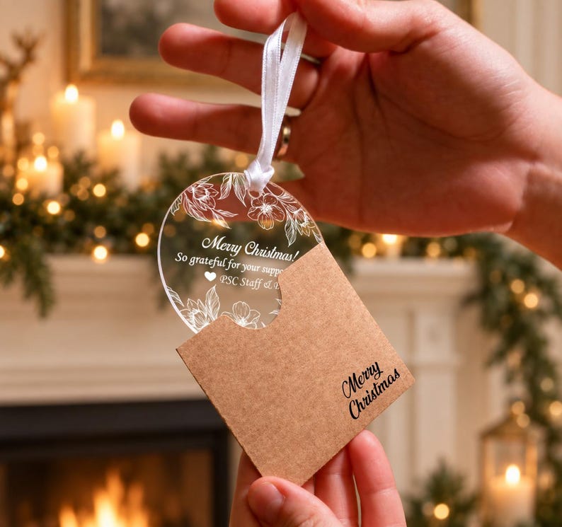 May include: A circular clear acrylic ornament featuring white line-art floral illustrations and the text Merry Christmas! So grateful for your support PSC Staff. It includes a white ribbon and a brown kraft paper sleeve with Merry Christmas printed.