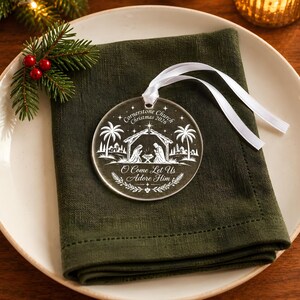 May include: A round, clear acrylic Christmas ornament featuring a white etched nativity scene with palm trees, stars, and the text "Cornerstone Church Christmas 2026" and "O Come Let Us Adore Him," finished with a white satin ribbon for hanging.