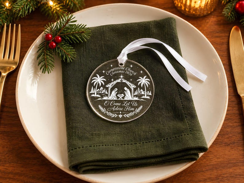 May include: A round, clear acrylic Christmas ornament featuring a white etched nativity scene with palm trees, stars, and the text "Cornerstone Church Christmas 2026" and "O Come Let Us Adore Him," finished with a white satin ribbon for hanging.