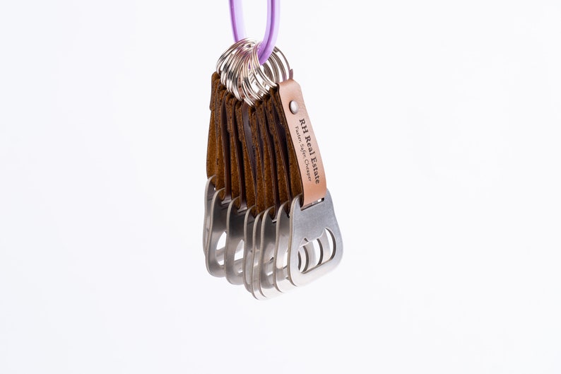 Another view of the leather-wrapped bottle opener keychains with brown leather wraps, hanging from a metal ring against a white background. The engraved text is clearly visible.