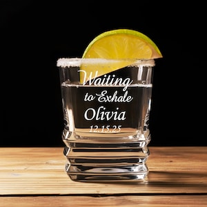 May include: A square-shaped glass filled with a clear liquid, rimmed with salt, and garnished with a lime wedge. The glass has the text "Waiting to Exhale Olivia 12.15.25" etched on the front. The glass sits on a wooden surface against a black background.