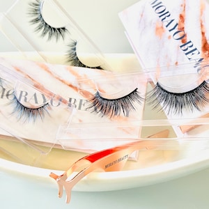 Op de afbeelding: Vier paar valse wimpers in doorzichtige plastic doosjes met een roze gouden rand. De doosjes liggen op een witte marmeren achtergrond. De tekst "MORAYO BEAUTY" is zichtbaar op de achtergrond. Een roze gouden wimperapplicator is ook zichtbaar.